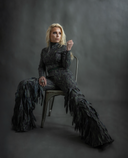 Stylish model in a dramatic feather-inspired outfit sitting elegantly on a chair against a textured backdrop.