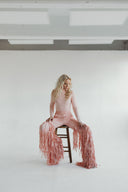 Lee Evans Lee in fringed pink Dancing Anyone outfit sitting on a wooden stool against a bright, minimalistic background.