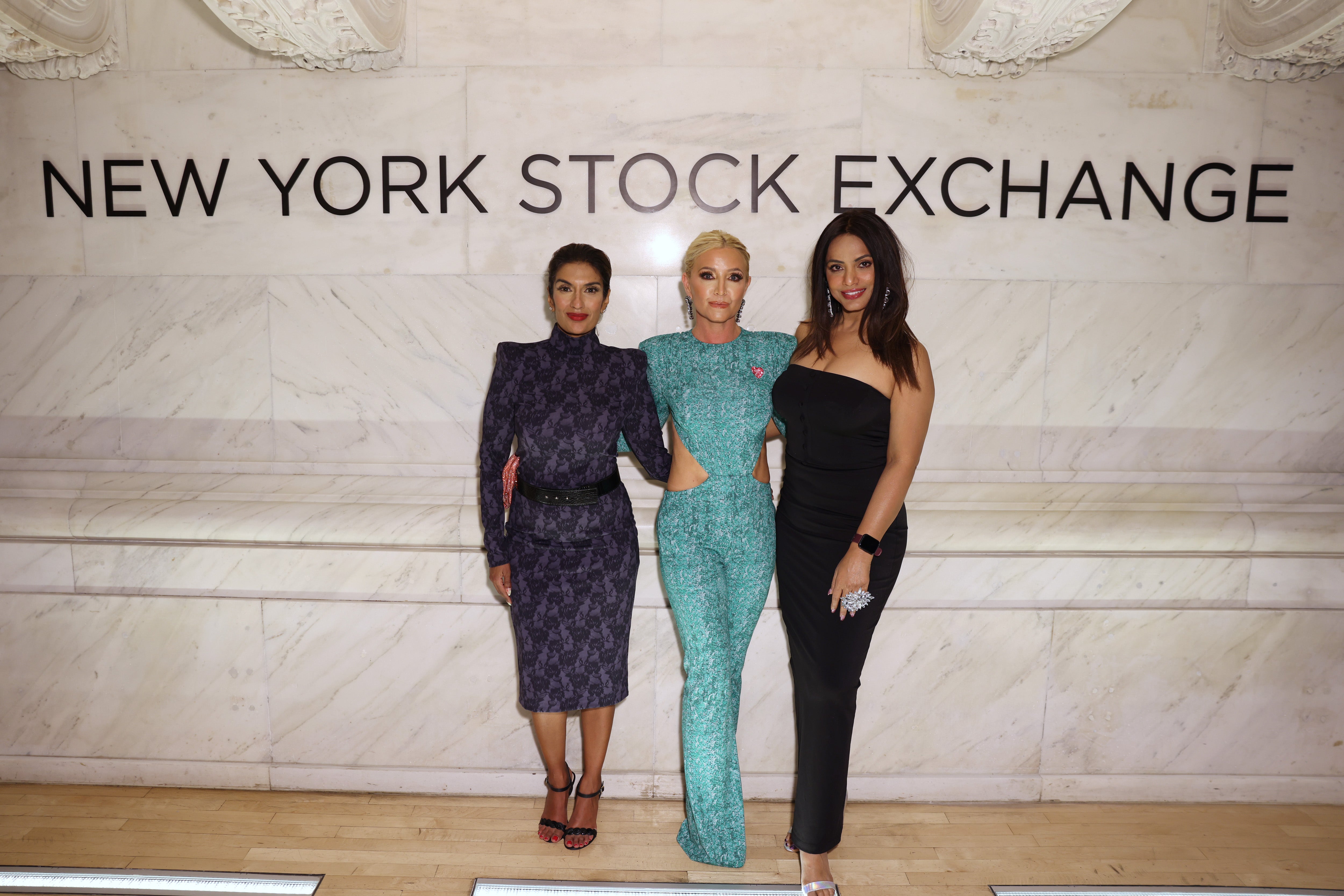 Three women posing in front of the New York Stock Exchange sign, showcasing fashion and elegance.