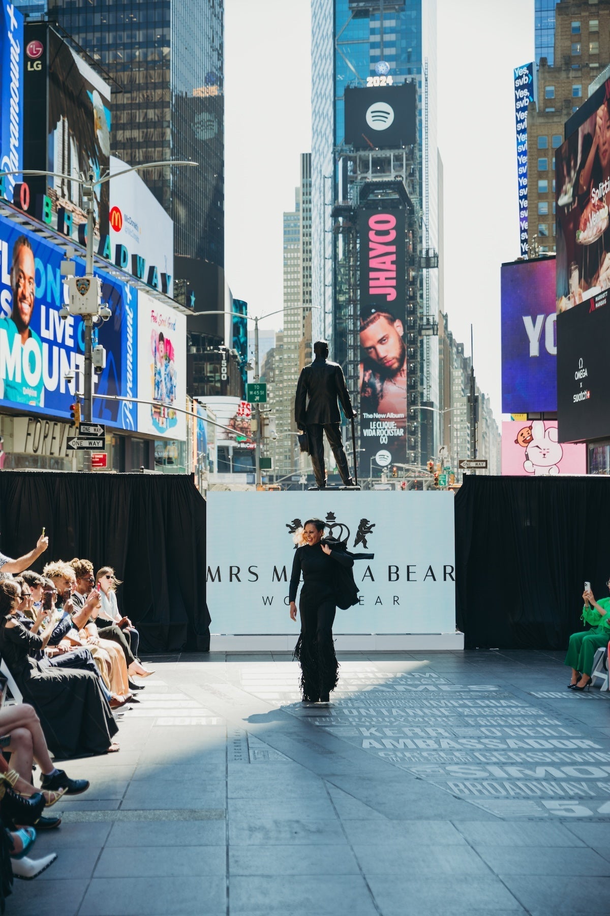 Model walking the runway at a fashion show in Times Square, surrounded by large digital billboards and an audience.