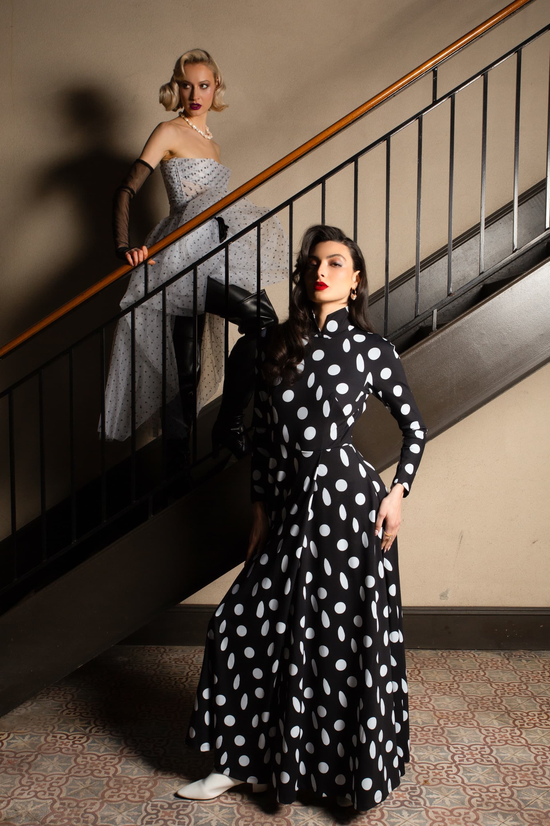 Two models on a staircase, one in a polka dot dress and the other in a strapless gown, showcasing bold fashion styling.