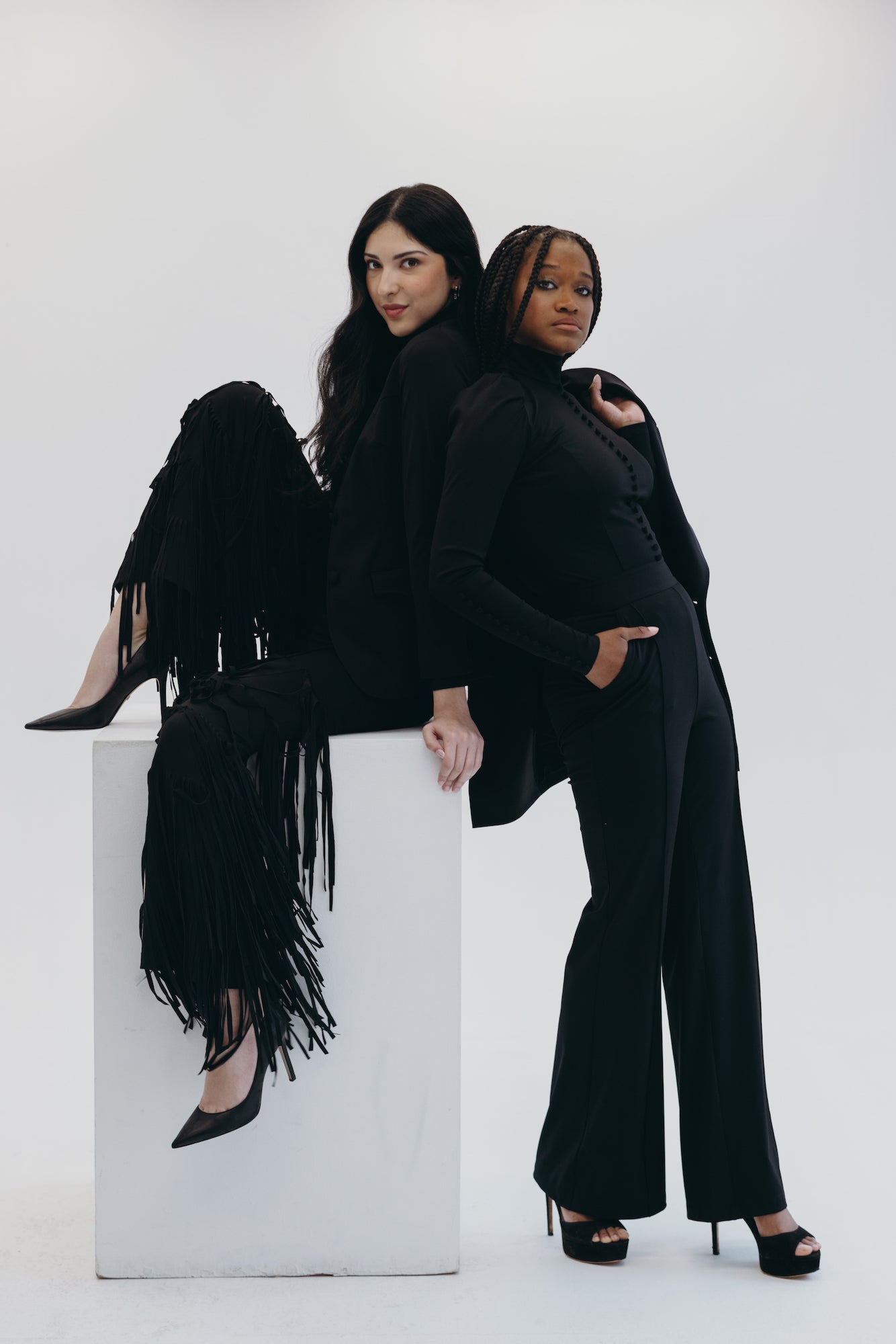 Two models in elegant black outfits pose creatively against a white background, showcasing modern fashion styles.