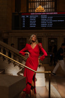 A model in a stunning red dress poses by the ticket vending area at a train station, with a departure board above.
