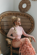 Woman in a pink top and fringed skirt sitting in a woven chair, showcasing a boho-chic interior style.