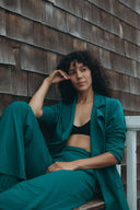 Woman in a teal Dahlia Blazer and matching pants, sitting casually against a wooden wall.