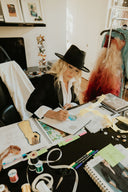 Woman in a black hat sketches designs at a cluttered workspace filled with fabric and sewing supplies.