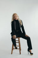 Confident woman in a black suit poses on a stool, showcasing a chic and professional style in a minimalist setting.