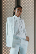 Elegant woman in a white suit with a bow tie, posing confidently against a patterned background.