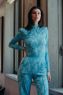 Model in blue floral jumpsuit posing by a window, showcasing modern fashion style and elegance.