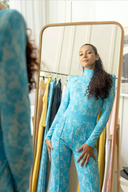 Woman in a blue patterned jumpsuit admiring herself in a mirror surrounded by stylish clothing.