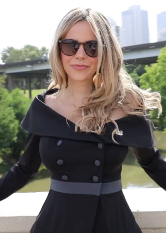 Young woman in sunglasses and an off-shoulder black dress poses outdoors with a green landscape and city skyline in background.