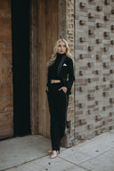 Stylish woman in a black Tea Time suit and turtleneck poses against a textured wall, showcasing modern fashion.