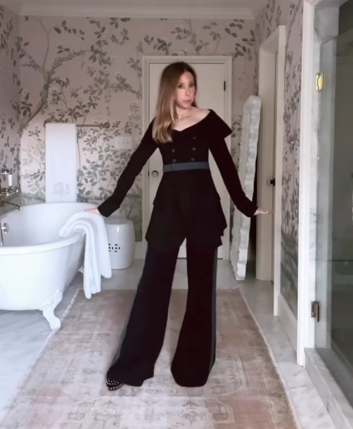 Woman in Tux All Day/All Night poses in a chic bathroom with floral wallpaper and a freestanding bathtub.
