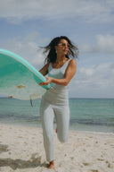 Woman in a light blue swimsuit carrying a surfboard on the beach with ocean waves in the background.