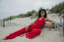 Woman in a stylish red jumpsuit lounging on the beach, wearing sunglasses with a sandy background and coastal grass.