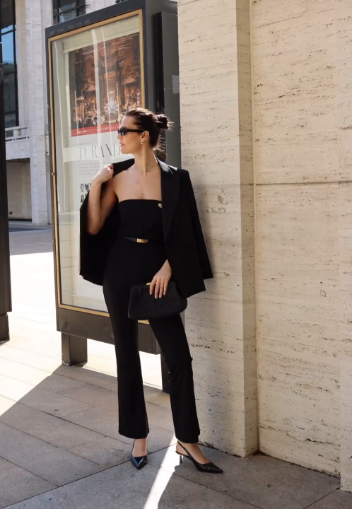 Elegant woman in Look at Me jumpsuit & blazer, accessorized with a clutch, posing stylishly outdoors.