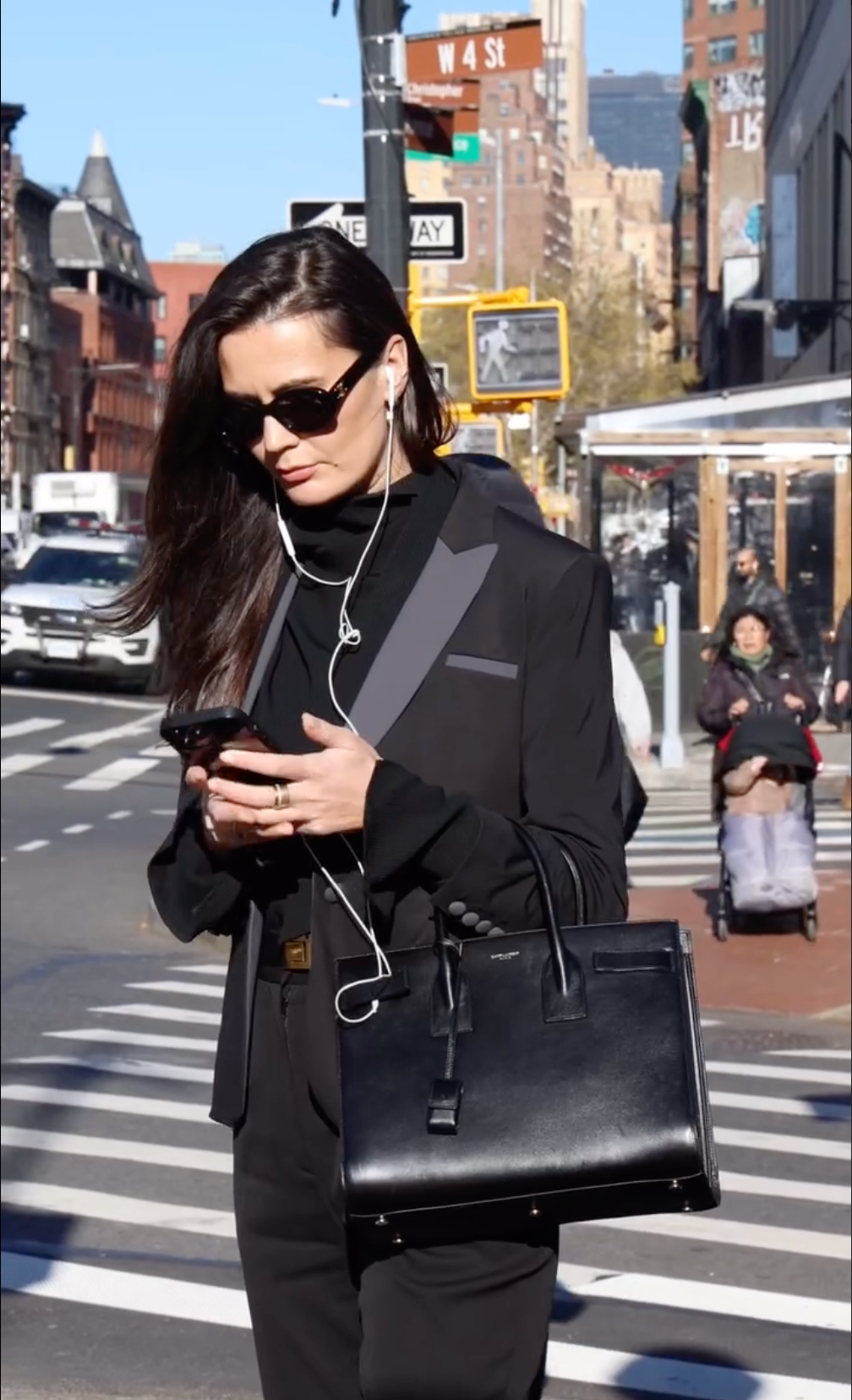 Woman in the Mad Hatter blazer and sunglasses walking in the city while using her phone, showcasing urban fashion style.