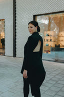 Woman in a stylish black outfit poses outdoors, showcasing a sleek open-back design against a modern retail backdrop.