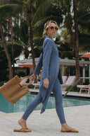 Trendy woman in blue outfit and sunglasses walking by a pool with a woven bag, surrounded by palm trees.