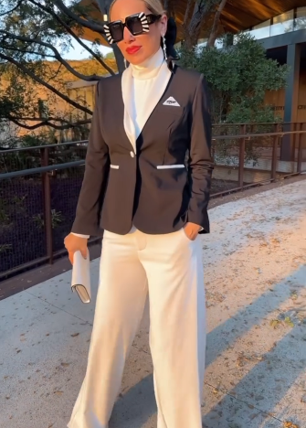 Stylish woman in Enza Marie blazer and white pants, wearing bold sunglasses, stands outdoors in modern surroundings.