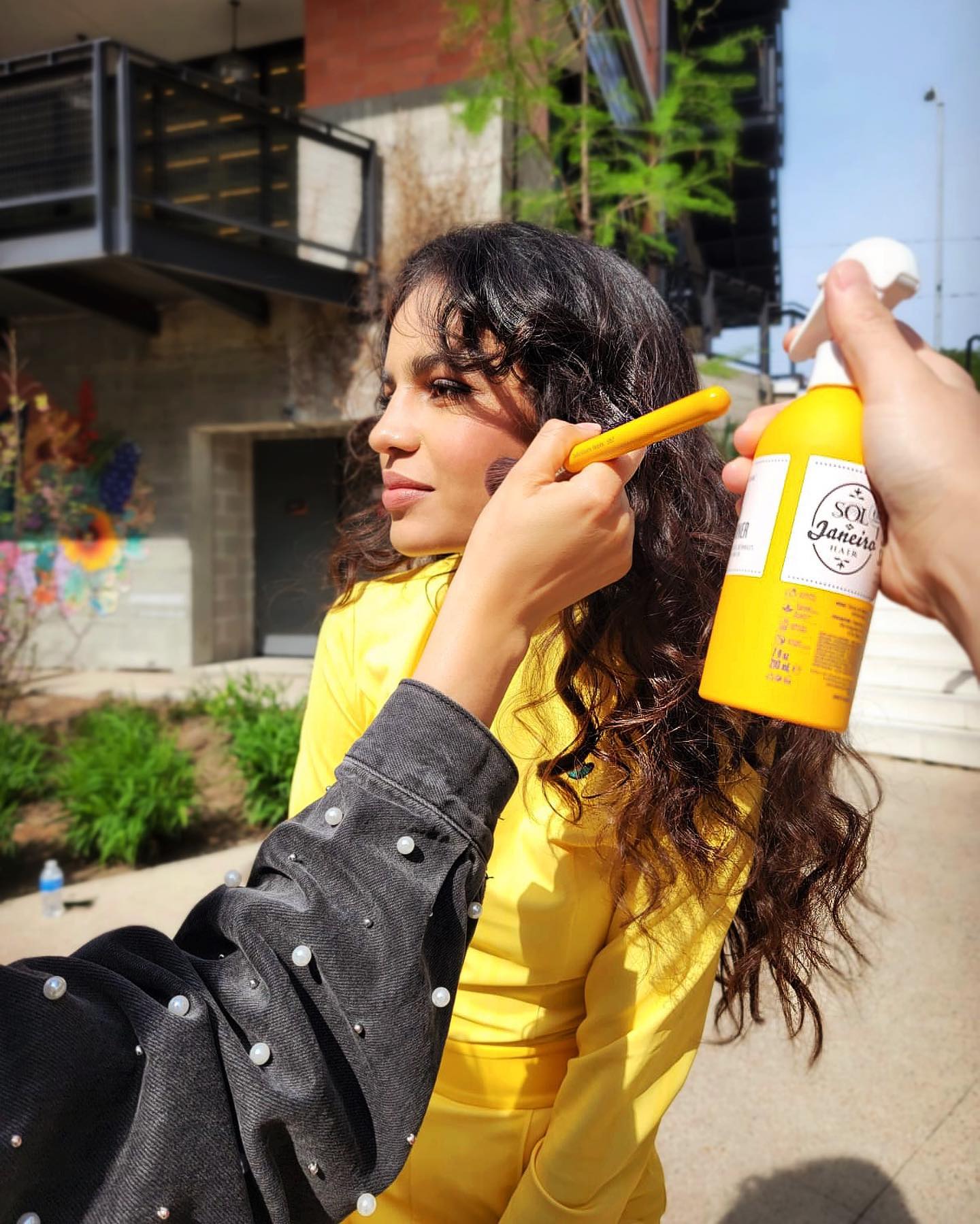 A woman with curly hair in a Milano being styled outdoors with a hair product and a styling tool.