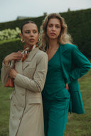 Two women in stylish suits posing outdoors, showcasing modern fashion trends in a lush green setting.