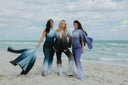 Three women in stylish gradient outfits pose on a beach, showcasing fashion against a scenic ocean backdrop.