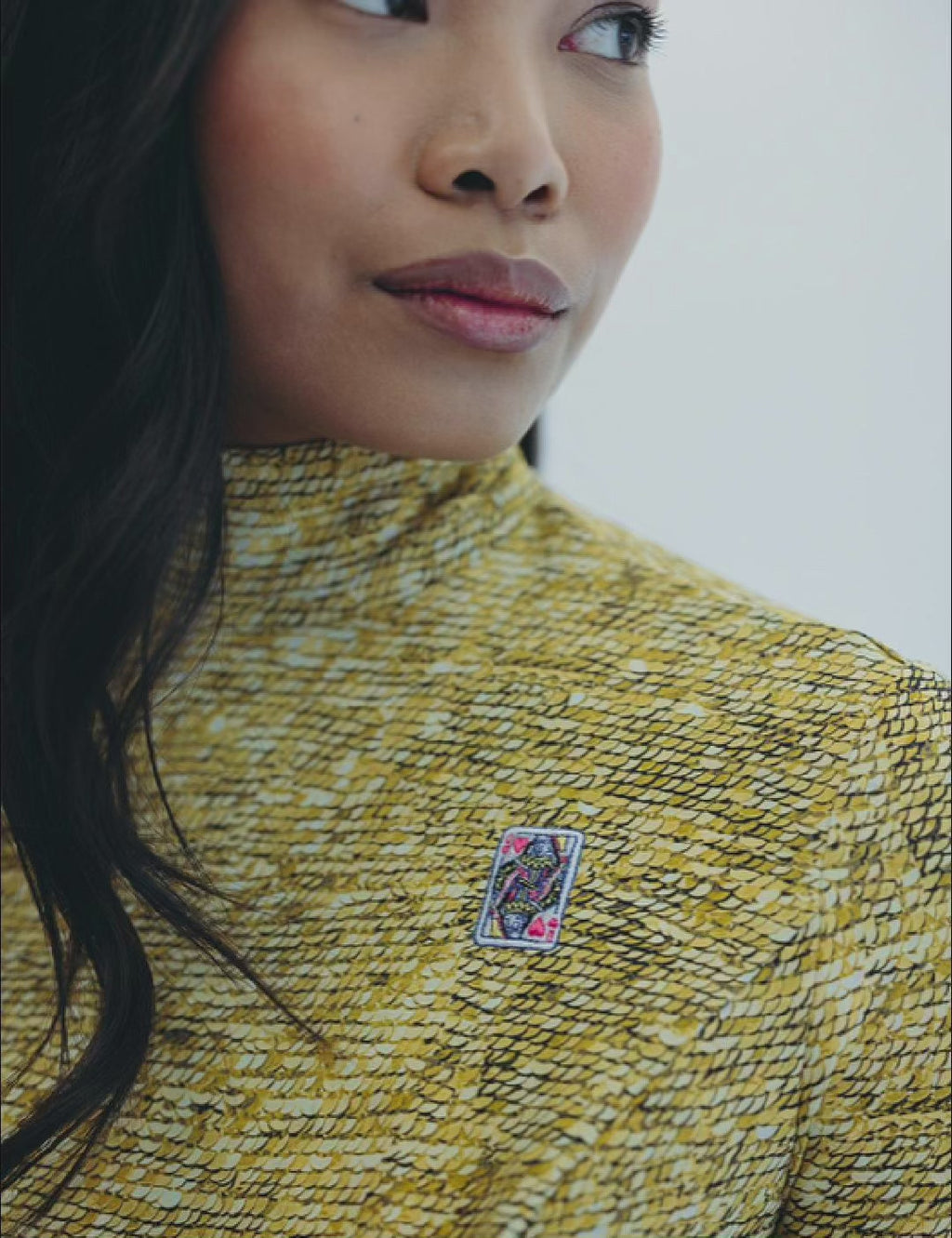 Close-up of a woman in a yellow sequin patterned 'Lucky Lady' jumpsuit featuring a decorative playing card pin on the collar.