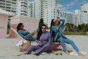 Three women in stylish beachwear relaxing on loungers near pastel pink beach huts, enjoying a sunny day.