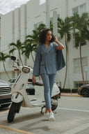 Woman in a stylish blue suit poses next to a scooter, surrounded by palm trees and urban scenery.