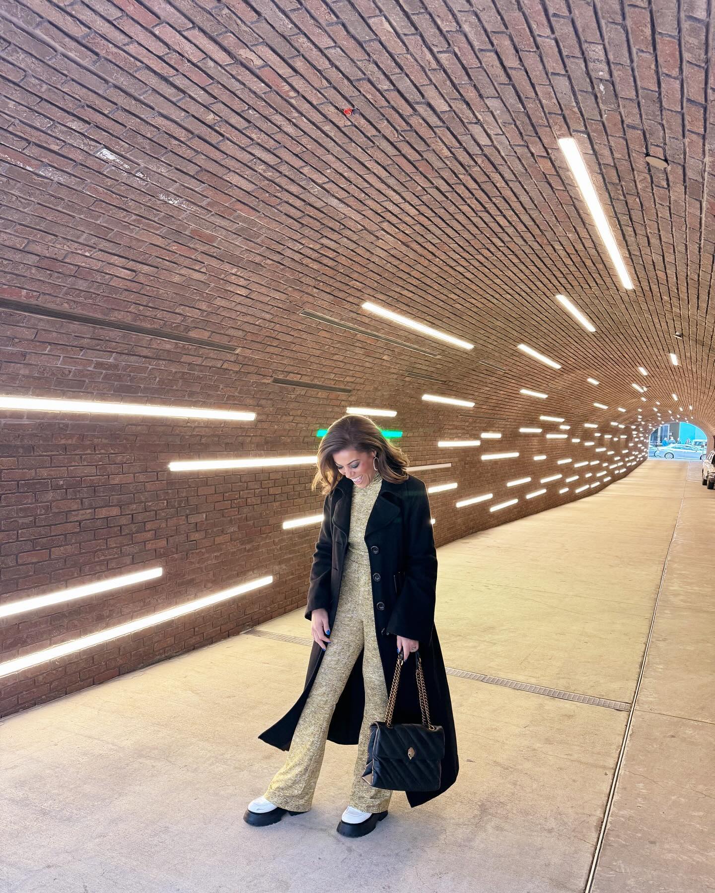 Fashionable woman in a stylish outfit walking through a modern, illuminated brick tunnel.