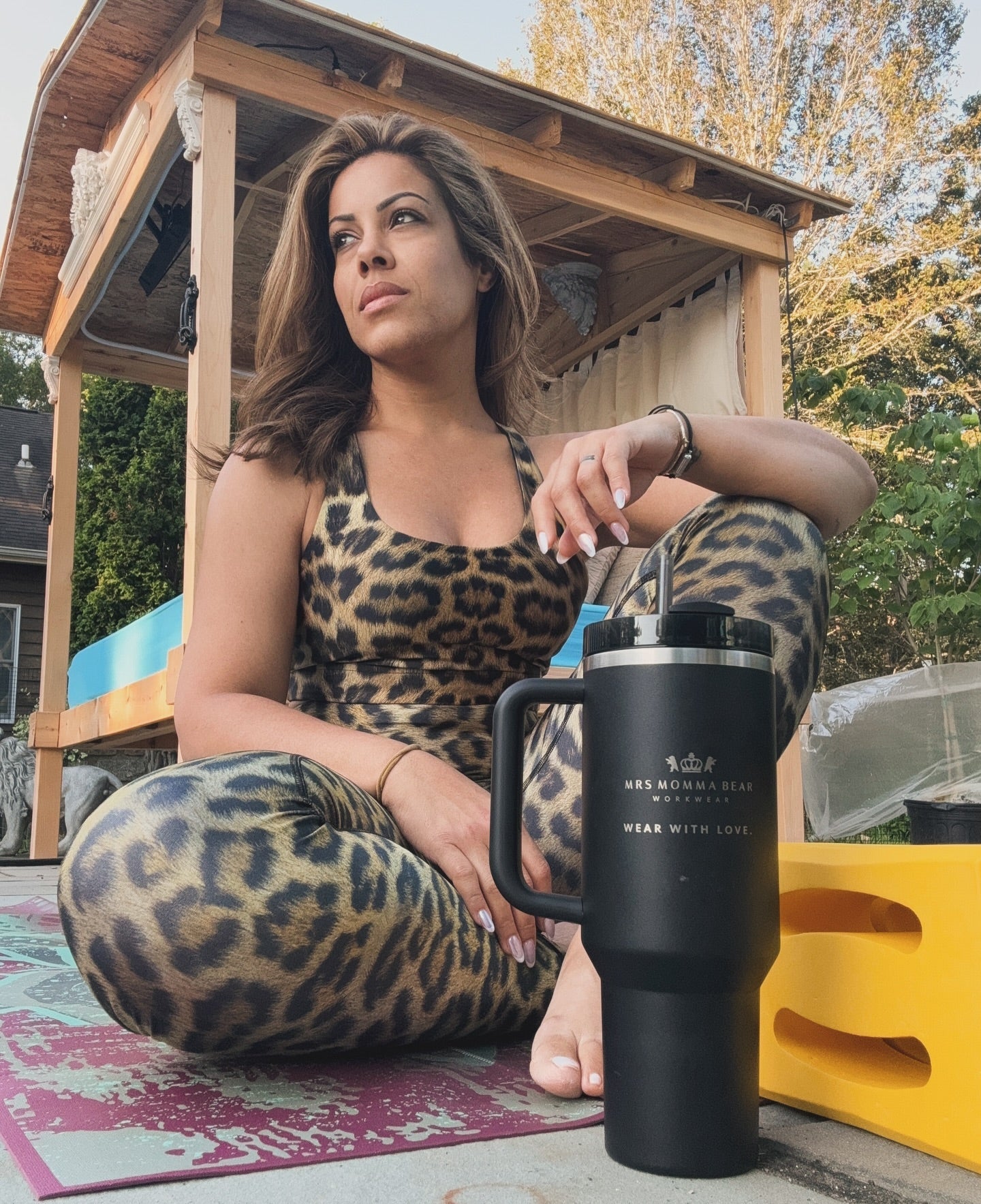 Woman in leopard print activewear sitting outdoors with a large black tumbler, enjoying a sunny day.